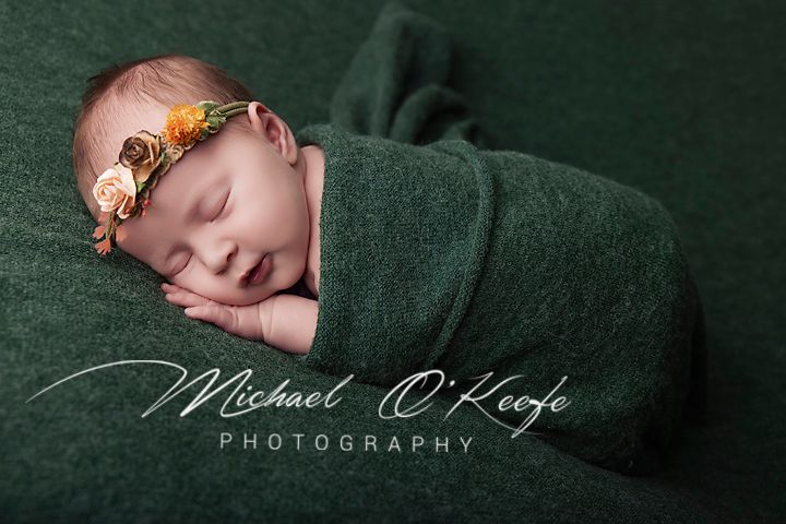 Newborn baby wrapped in green swaddle with floral headband, sleeping peacefully in professional photography portrait.