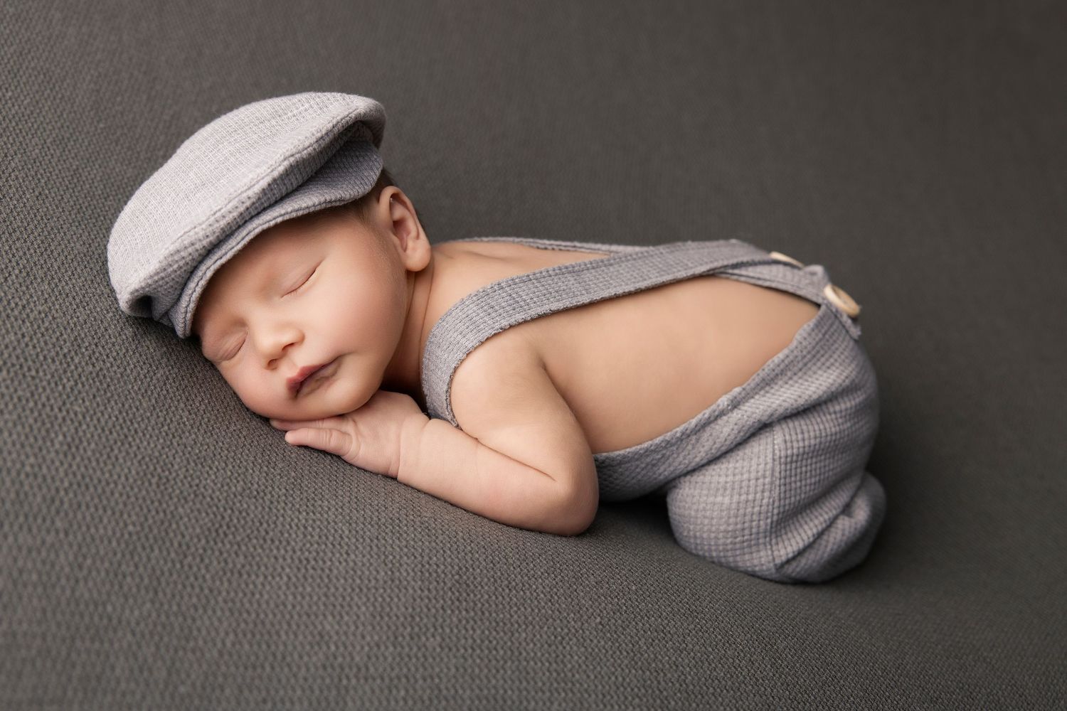Sleeping newborn in gray newsboy cap and suspenders on dark backdrop.