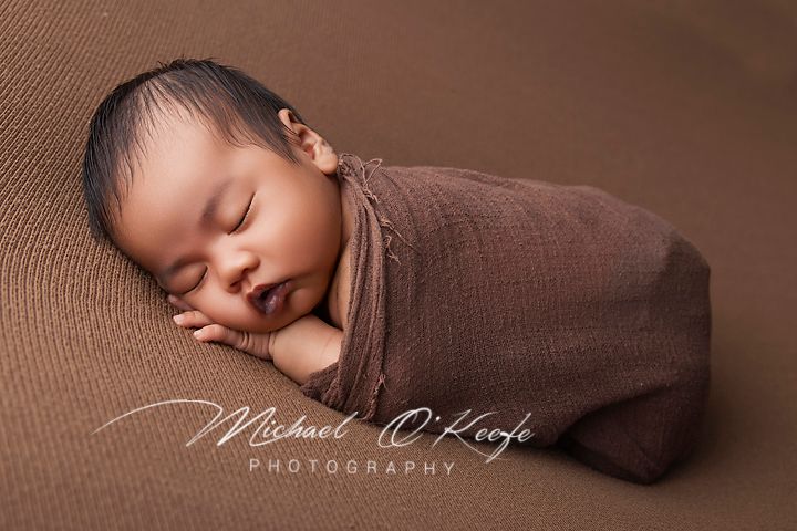 Newborn baby swaddled in brown wrap sleeping peacefully on brown backdrop by Michael O'Keefe Photography.