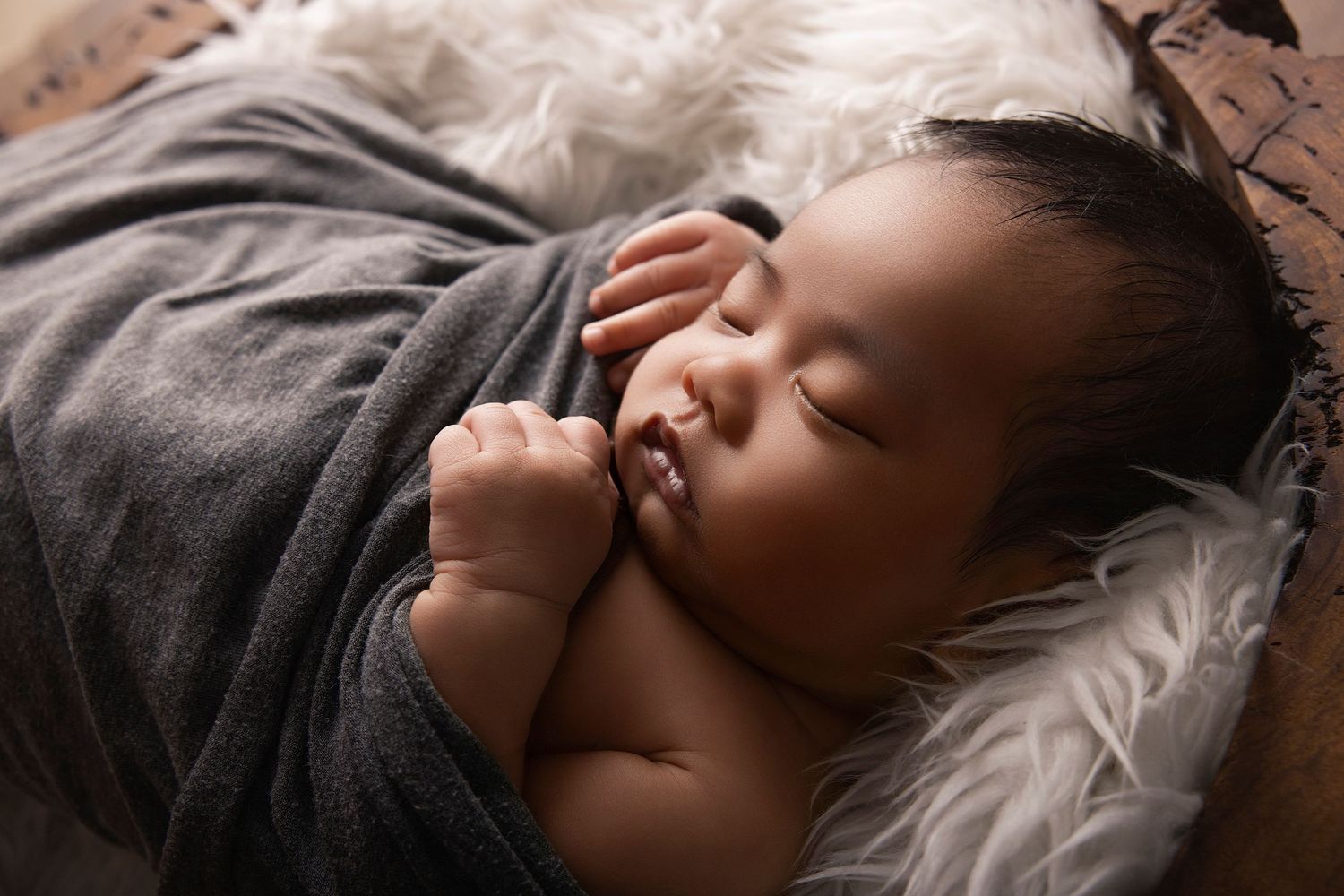 Peaceful newborn baby sleeping wrapped in a soft gray swaddle blanket inside a cozy wooden bowl.