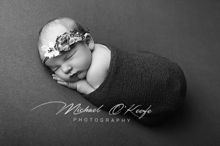 Black and white newborn photography of a sleeping baby wrapped in a dark swaddle with a floral headband by Michael O'Keefe.