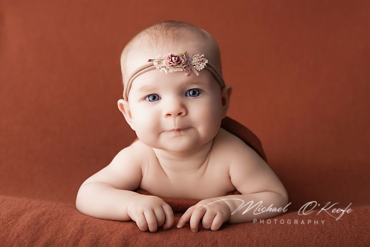 A cute baby with a floral headband poses on a rust-colored blanket in a professional studio portrait photo.