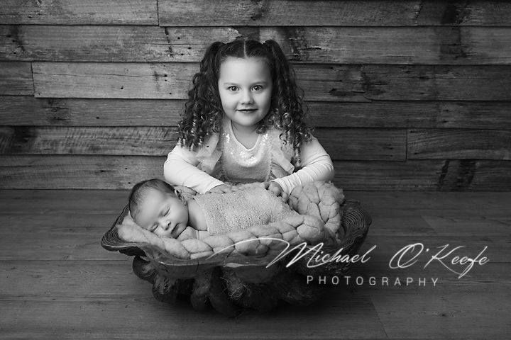 Black and white photo of a young girl with curly hair holding a sleeping newborn baby in a wooden bowl prop.