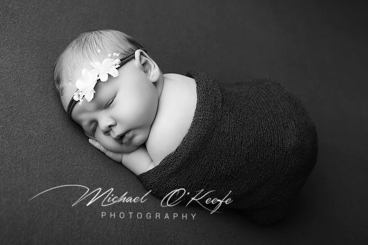 Black and white newborn photo of a sleeping baby wrapped in a dark knit blanket wearing a delicate floral headband.