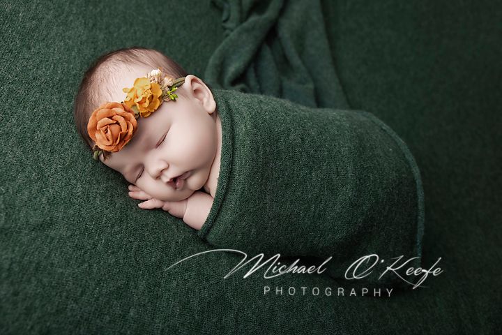 Newborn baby swaddled in green wrap with orange floral headband, sleeping peacefully in Michael O'Keefe Photography portrait.