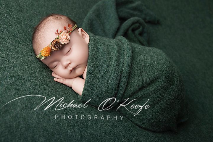 Newborn baby swaddled in green wrap with floral headband, sleeping peacefully in Michael O'Keefe Photography portrait.