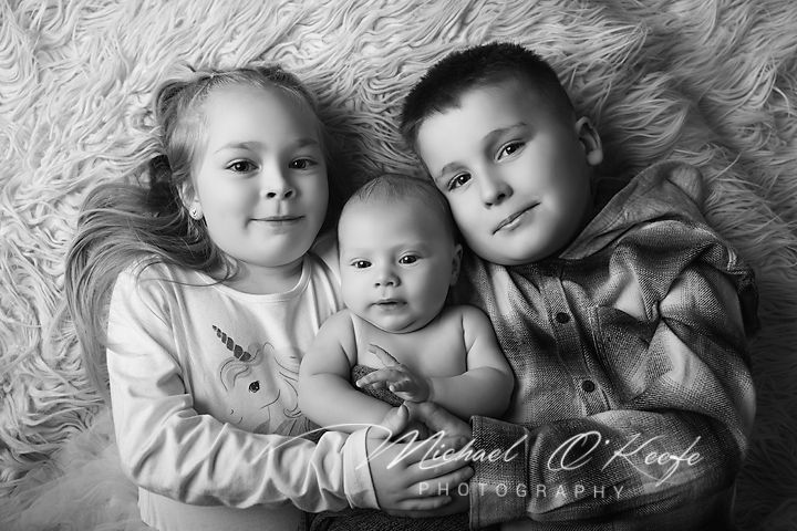 Black and white photo of three siblings, two older children and a newborn baby, lying together on a fluffy rug.
