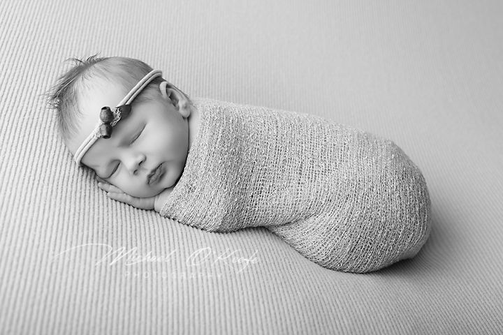 Black and white photo of a sleeping newborn baby swaddled in a soft wrap, wearing a delicate headband.