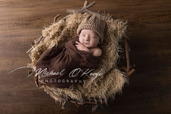 Newborn baby sleeping peacefully in a rustic nest bowl with soft fur, wearing a brown knit hat, by Michael O'Keefe Photography.