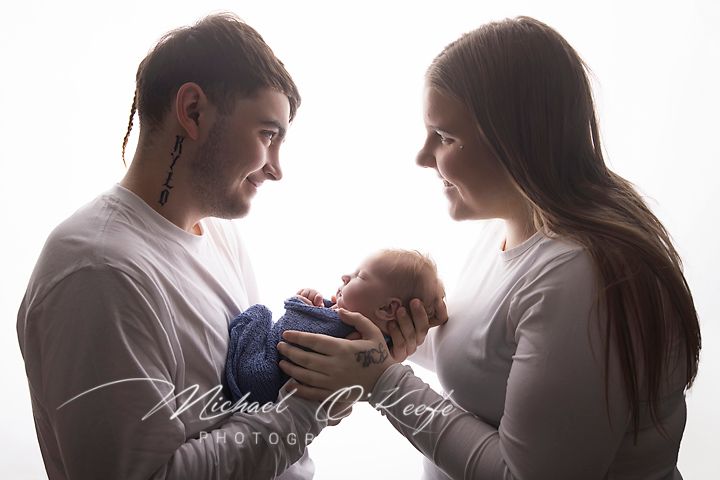 New parents lovingly cradle their sleeping newborn baby in a bright white studio portrait photo.