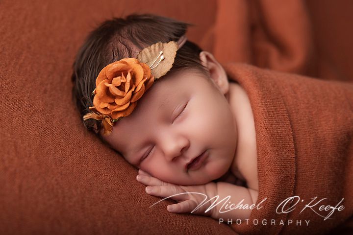 Newborn baby sleeping peacefully wrapped in burnt orange fabric with a floral headband, professional photography.