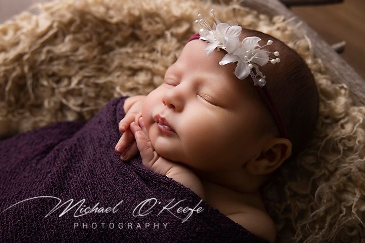 Sleeping newborn baby girl wrapped in purple swaddle with white floral headband, professional newborn photography.