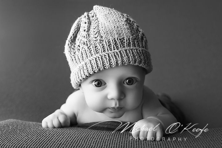 Black and white photo of an adorable baby wearing a knit hat, lying on stomach with big expressive eyes.
