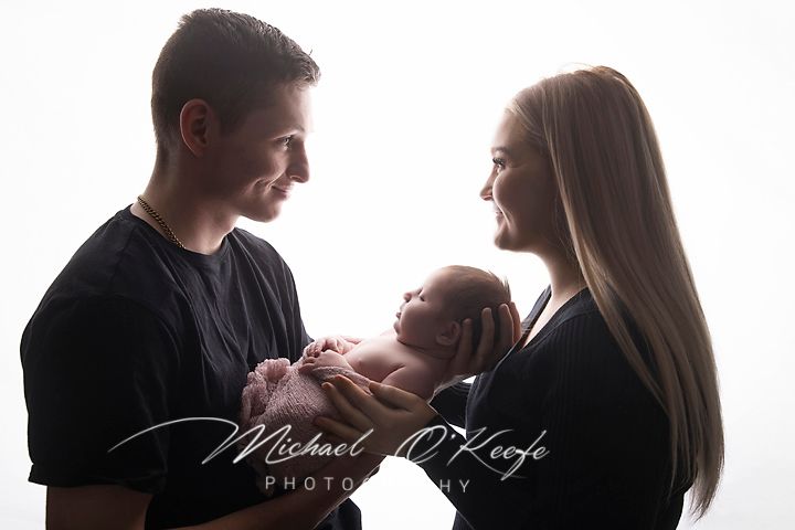 Silhouetted new parents smiling at their sleeping newborn baby cradled in their hands in a bright, high-key newborn photography portrait.