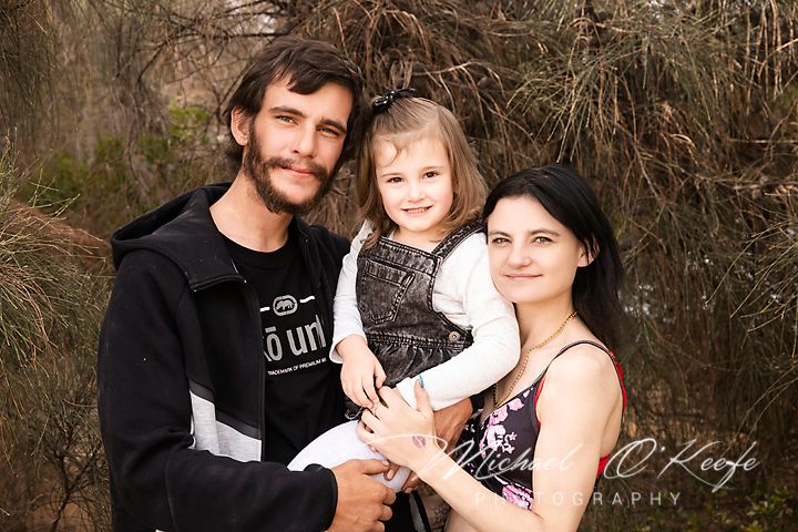 A family poses together outdoors in a natural setting with brown foliage in the background.