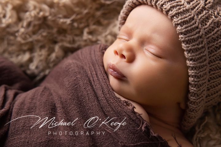 Sleeping newborn baby swaddled in brown fabric lying on textured blanket during professional photo session.