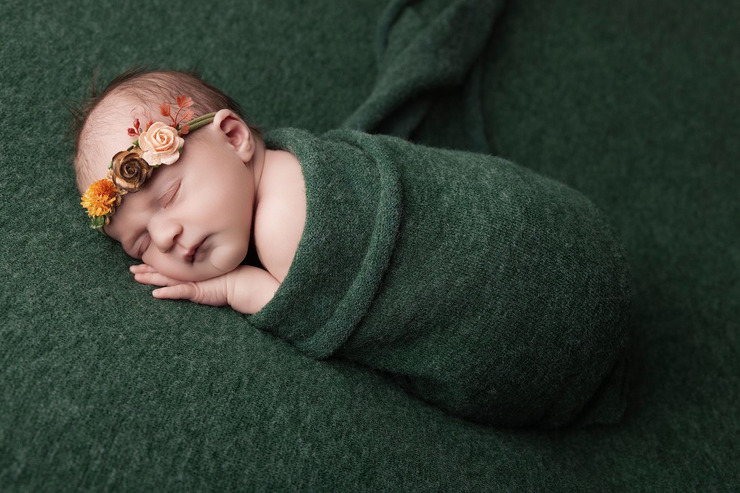 Newborn baby swaddled in green wrap with floral headband, sleeping peacefully on a dark green backdrop.
