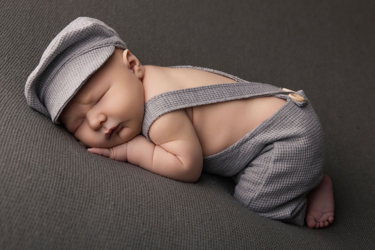 Sleeping newborn baby wearing gray knit suspender outfit and matching newsboy cap on gray background.