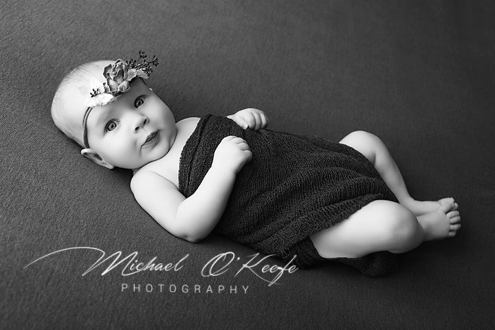Black and white photo of a cute baby in a knit romper and floral headband, lying on a gray backdrop.