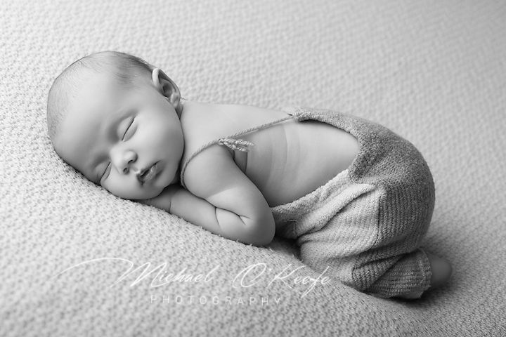 Black and white newborn photo of a sleeping baby in knit overalls, curled up peacefully on a soft blanket.
