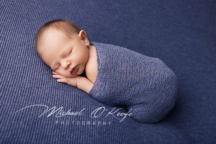 Newborn baby swaddled in blue wrap sleeping peacefully on blue textured backdrop by Michael O'Keefe Photography.