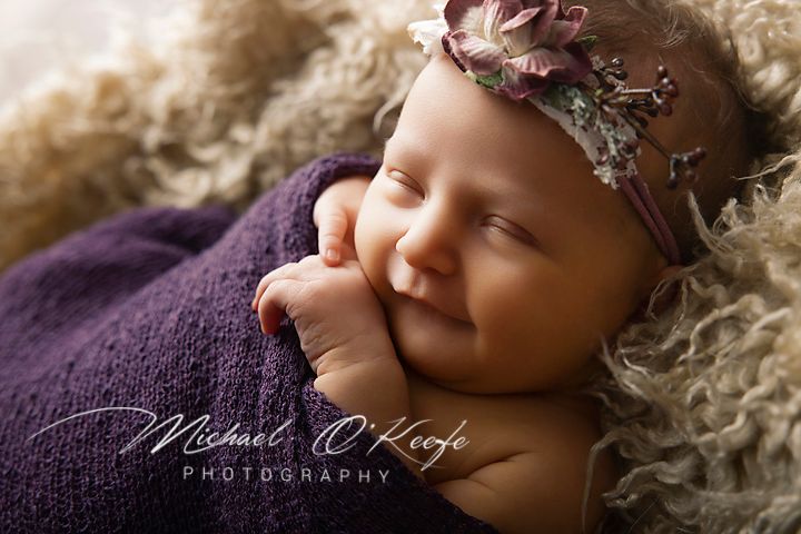 Newborn baby girl sleeping peacefully wrapped in purple, wearing a floral headband, photographed by Michael O'Keefe Photography.