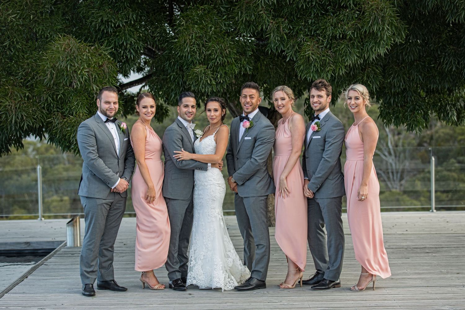 Wedding party in gray suits and pink dresses poses together on a wooden deck.