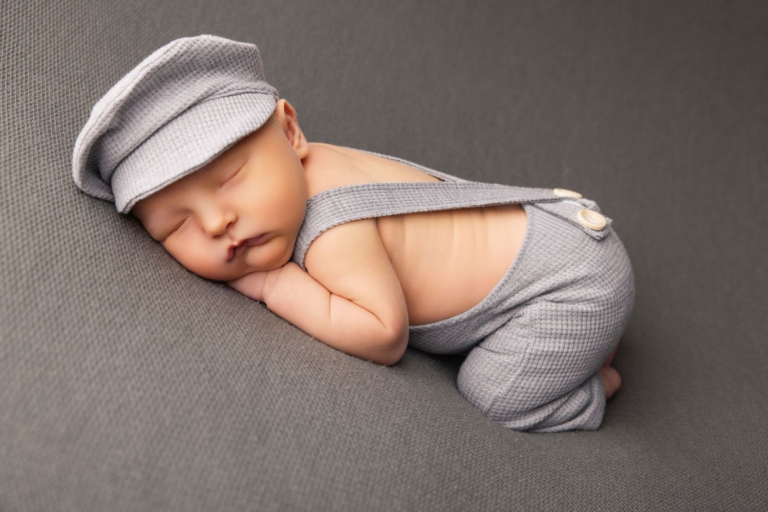 Sleeping newborn wearing gray newsboy cap and suspenders on dark backdrop.