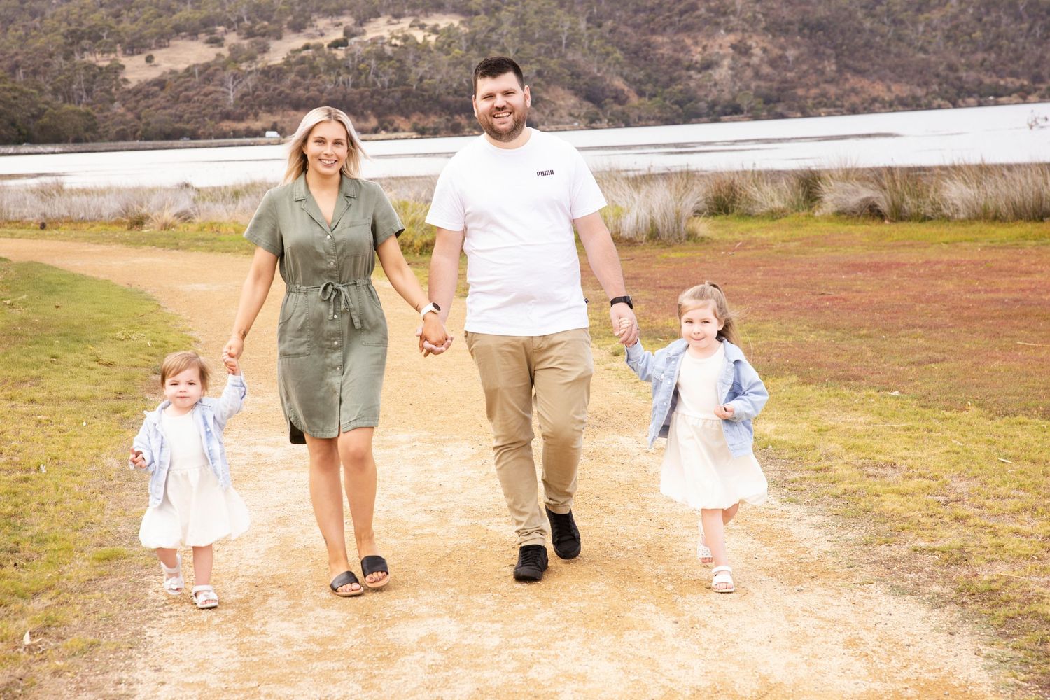 Family enjoys a playful walk together on a dirt trail surrounded by tall grass and trees.