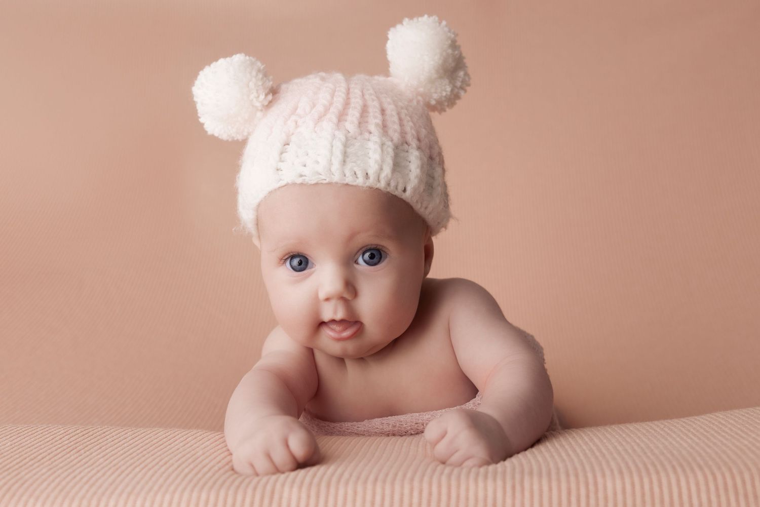 Baby wearing white knit hat with pom poms lying on pink background.