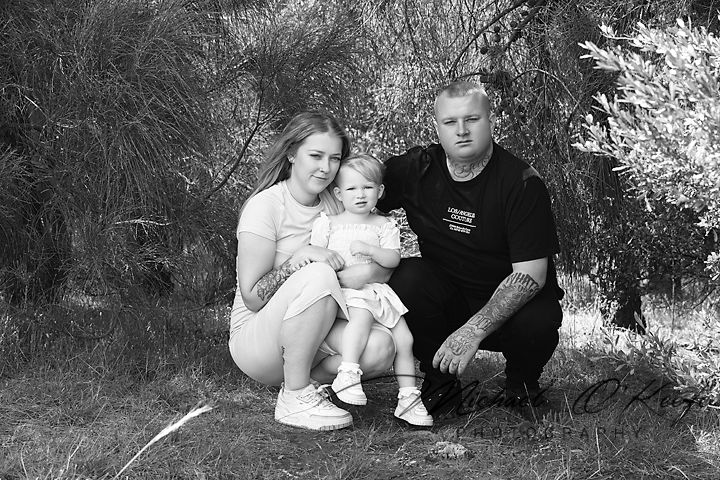 A black and white portrait of a family of three sitting together outdoors against a natural backdrop.