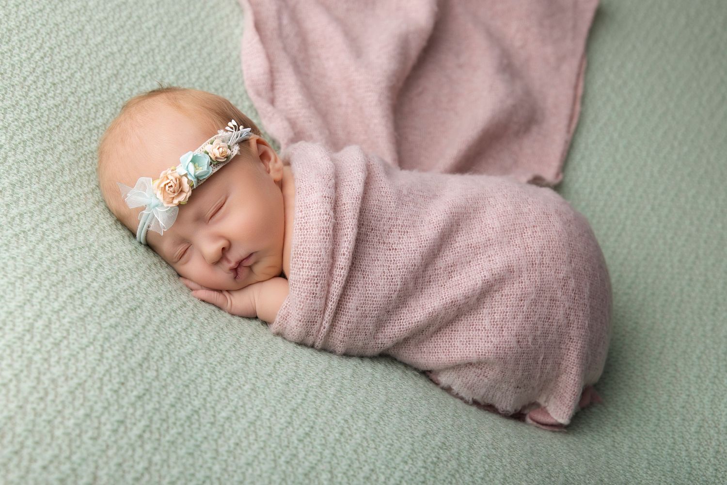 Newborn baby girl swaddled in pink wrap with floral headband sleeping peacefully on mint green blanket.