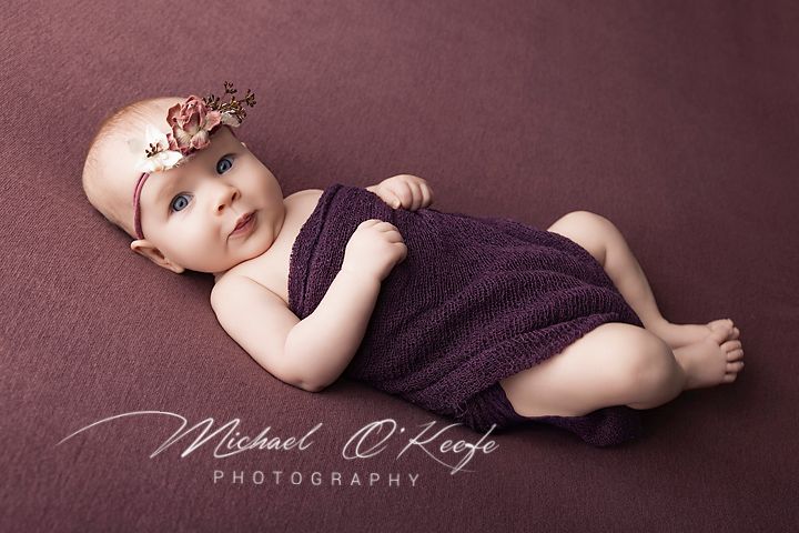 A baby girl in a purple romper and floral headband poses on a mauve backdrop in a professional newborn photo.