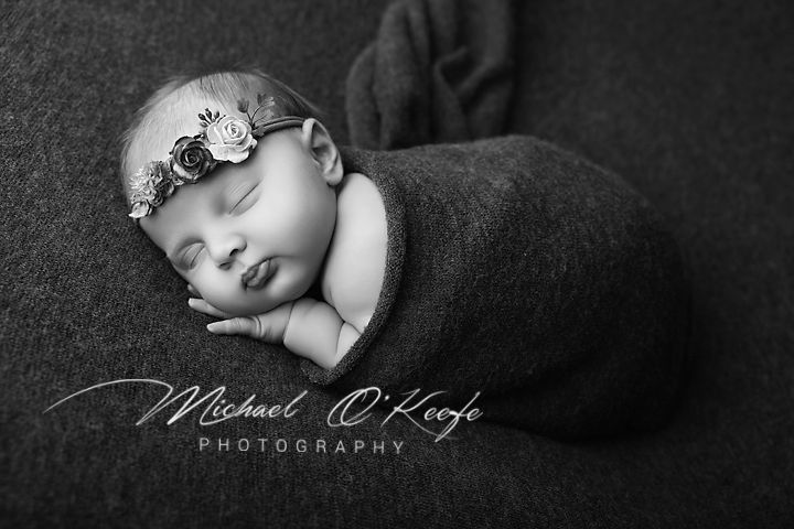 Black and white newborn portrait of a swaddled baby wearing a floral headband, sleeping peacefully by Michael O'Keefe Photography.