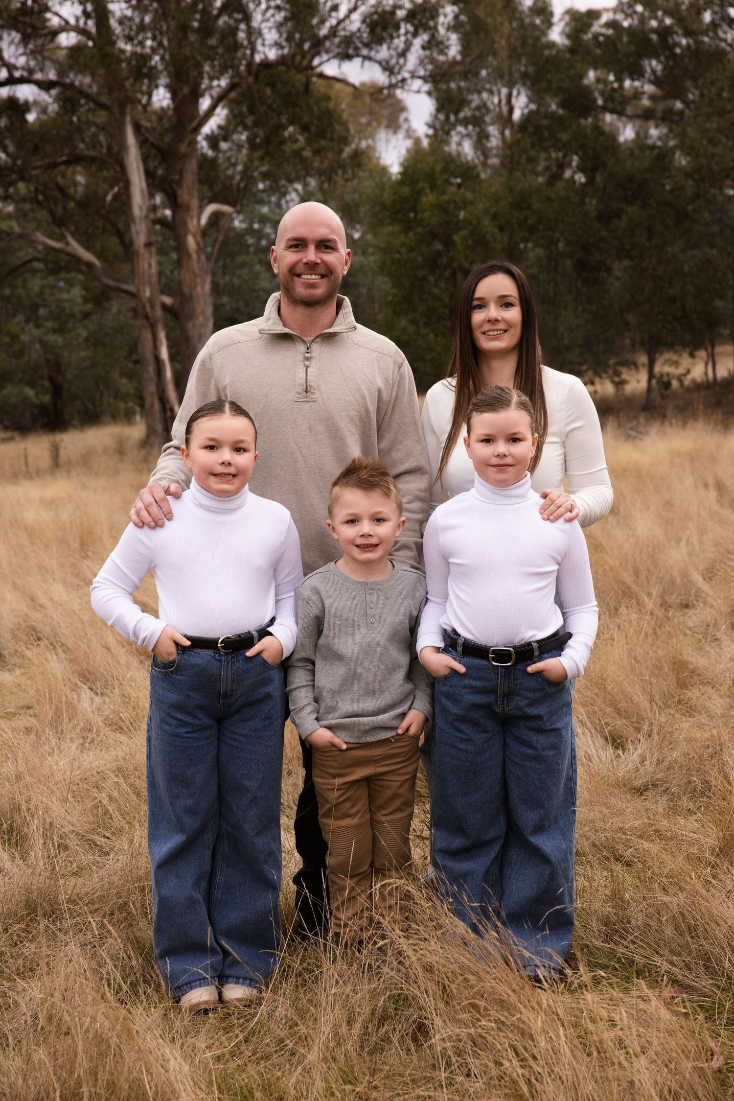 Family poses together in casual attire against a backdrop of tall grass and trees.
