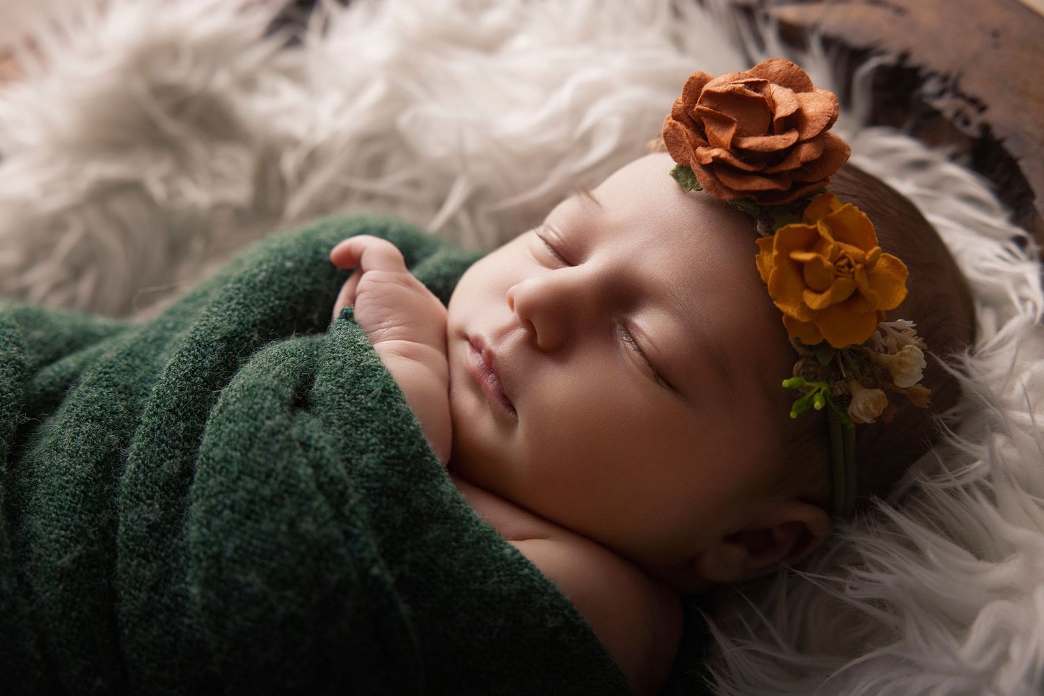 Sleeping newborn baby wrapped in green knit swaddle with orange floral headband on fluffy white fur backdrop.