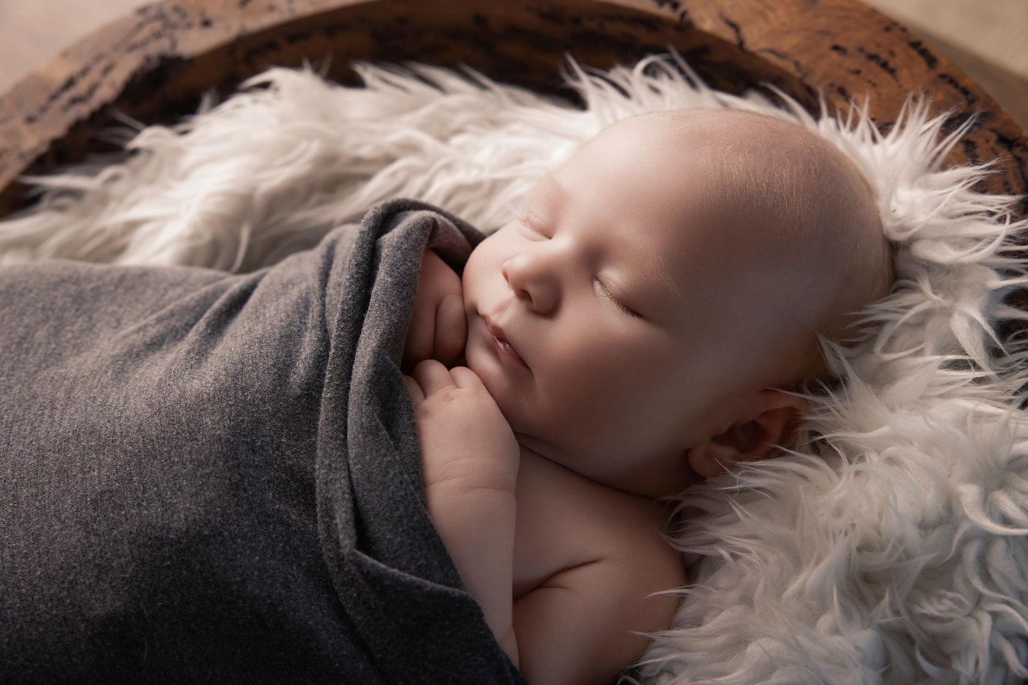 Newborn baby sleeping peacefully wrapped in gray blanket on fluffy white fur inside a wooden bowl.