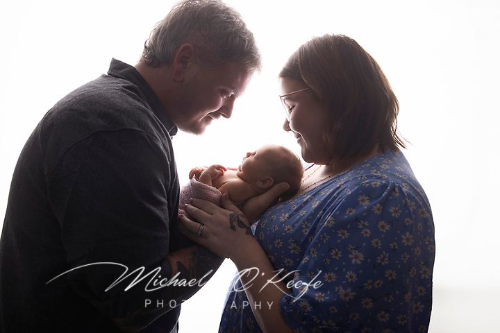 New parents tenderly cradle their sleeping newborn baby in a beautifully backlit studio portrait by Michael O'Keefe Photography.