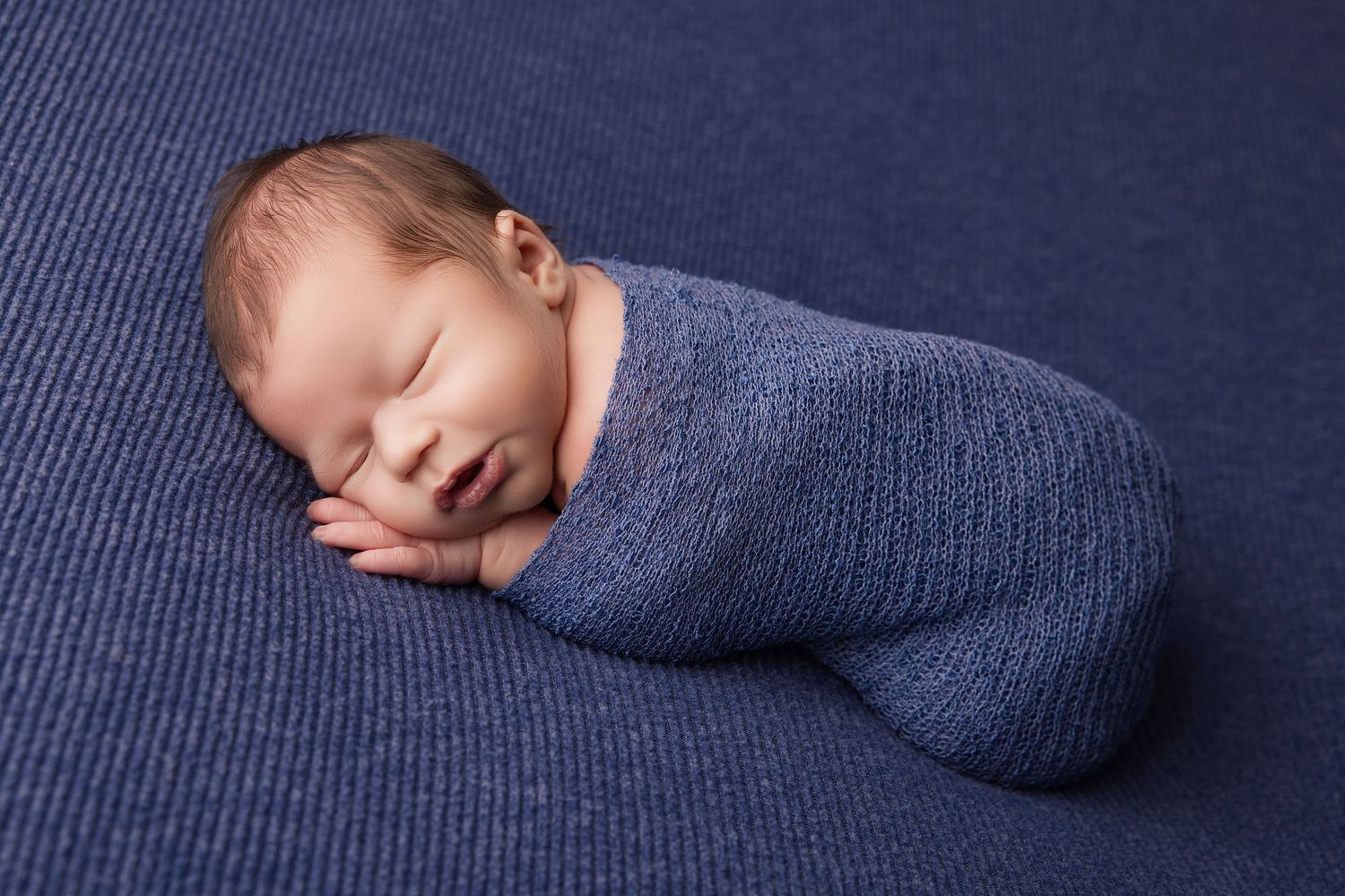 A sleeping newborn baby wrapped in a soft blue swaddle blanket on a navy blue textured background.