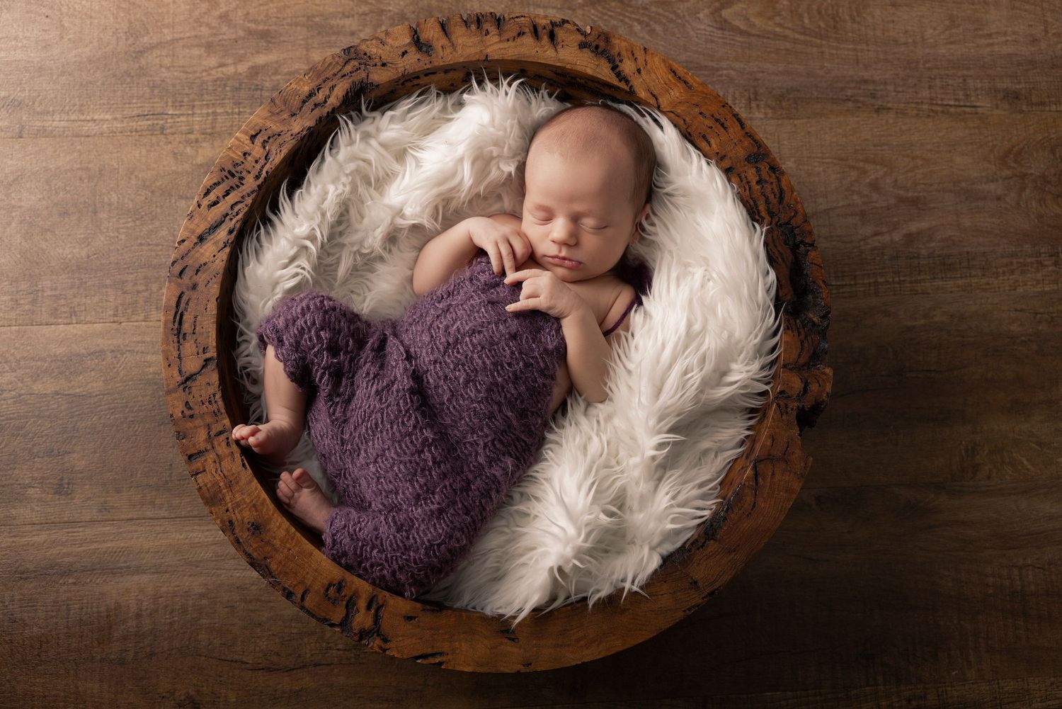 Newborn sleeping peacefully in wooden bowl lined with white fur and purple wrap.