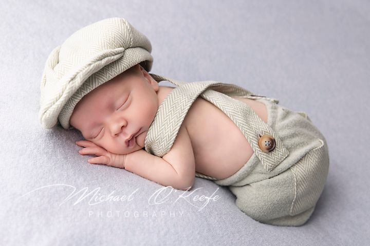 A peaceful sleeping newborn wearing a light beige knit outfit lies curled up with soft blankets in a studio setting.