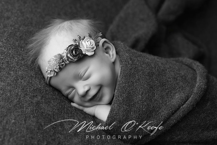 Black and white portrait of a sleeping newborn wearing a delicate floral headband on a dark blanket.