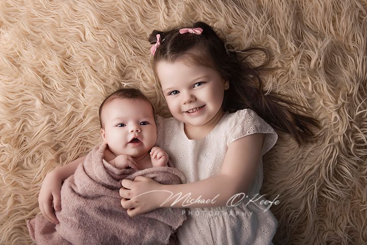 A smiling toddler girl in a white dress cuddles a newborn baby wrapped in pink on a fluffy beige fur rug.