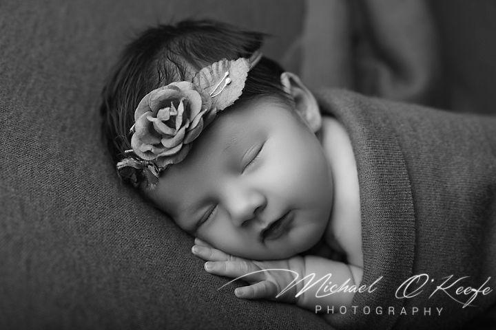 Black and white newborn photo of sleeping baby girl wearing floral headband, wrapped in soft blanket.