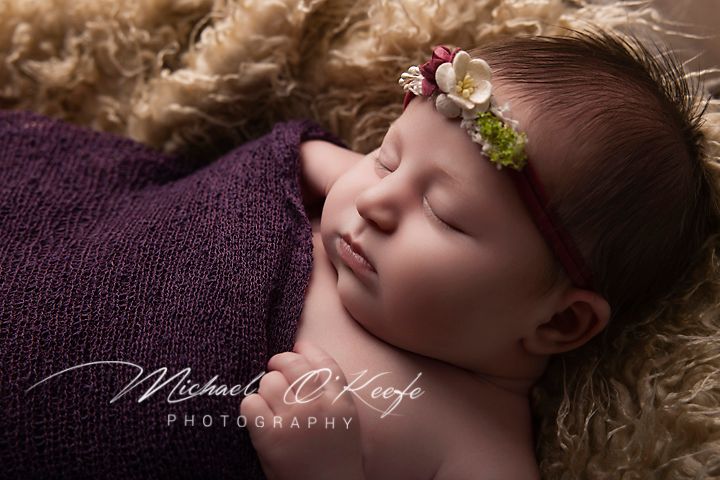 Sleeping newborn baby girl with a floral headband swaddled in a purple wrap on a cream fluffy blanket in a professional newborn photo.
