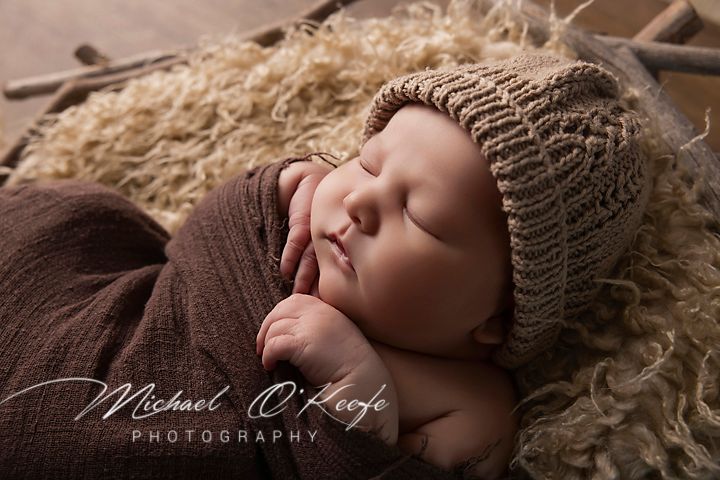 Sleeping newborn wrapped in brown swaddle wearing knit beanie rests peacefully on soft furry blanket.