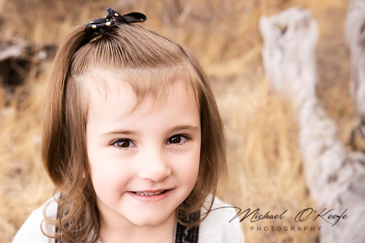 A young child in a brown sweater poses for a portrait outdoors with a natural blurred background.