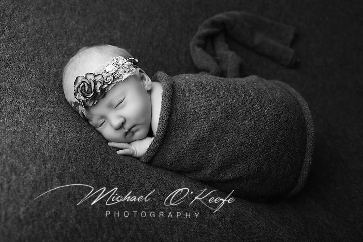 Black and white photo of a sleeping newborn baby swaddled in a gray wrap with a floral headband.