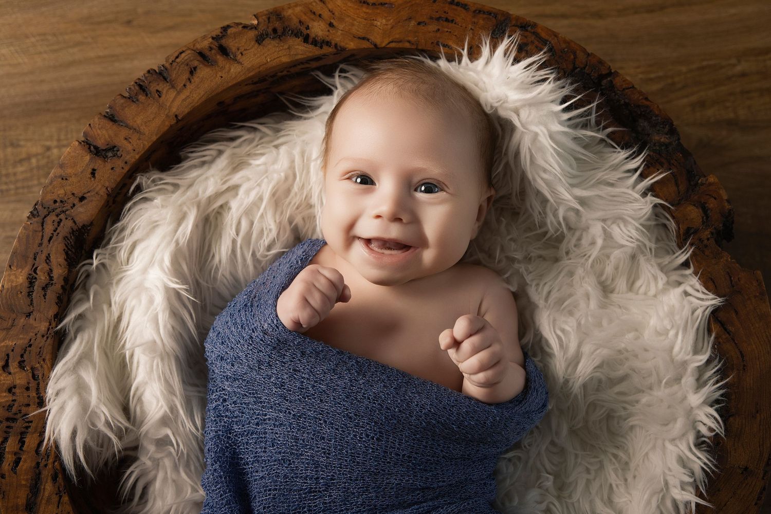 Smiling newborn baby wrapped in blue swaddle nestled in fluffy white fur inside a rustic wooden bowl.