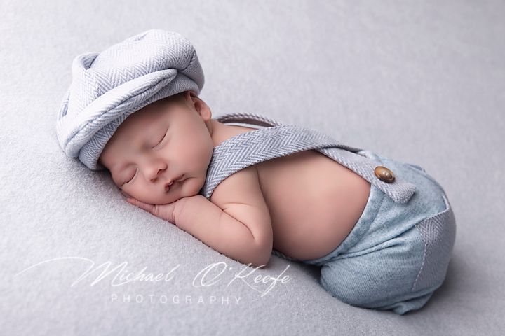 Sleeping newborn baby wearing a light blue knit cap and suspender shorts on a soft gray backdrop.